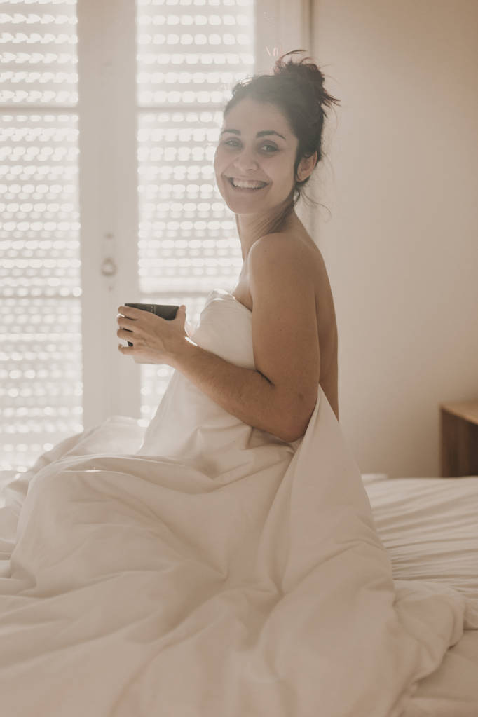 mujer desnuda cubierta de sábanas blancas sentada en la cama y disfrutando de una taza de café. el sol está entrando por la ventana. Estilo de vida interior
 - Foto, Imagen