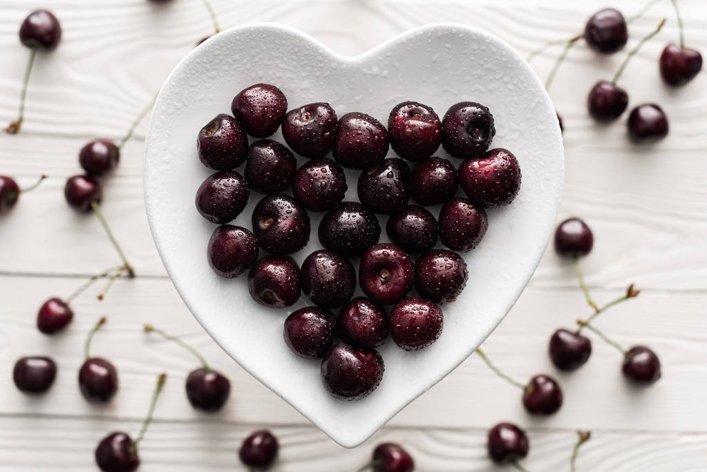 top view of fresh, sweet and ripe cherries covered with water drops on heart shaped plate  - Photo, Image