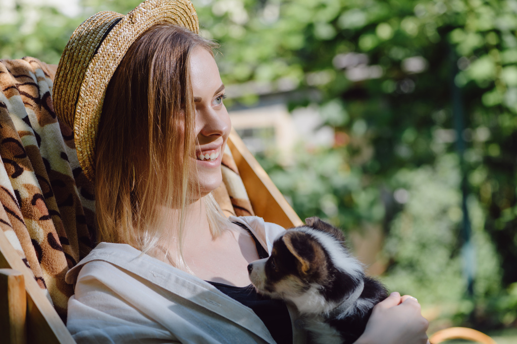 Gelukkig blond meisje in stro hoed houden Corgi puppy terwijl zittend in dek stoel in de tuin - Foto, afbeelding