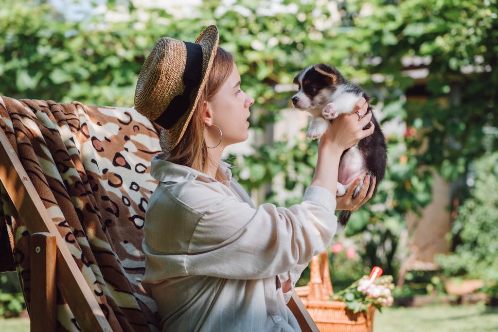 Widok z boku blond dziewczyna w słomy kapelusz trzyma szczeniaka siedząc w leżaku w ogrodzie - Zdjęcie, obraz