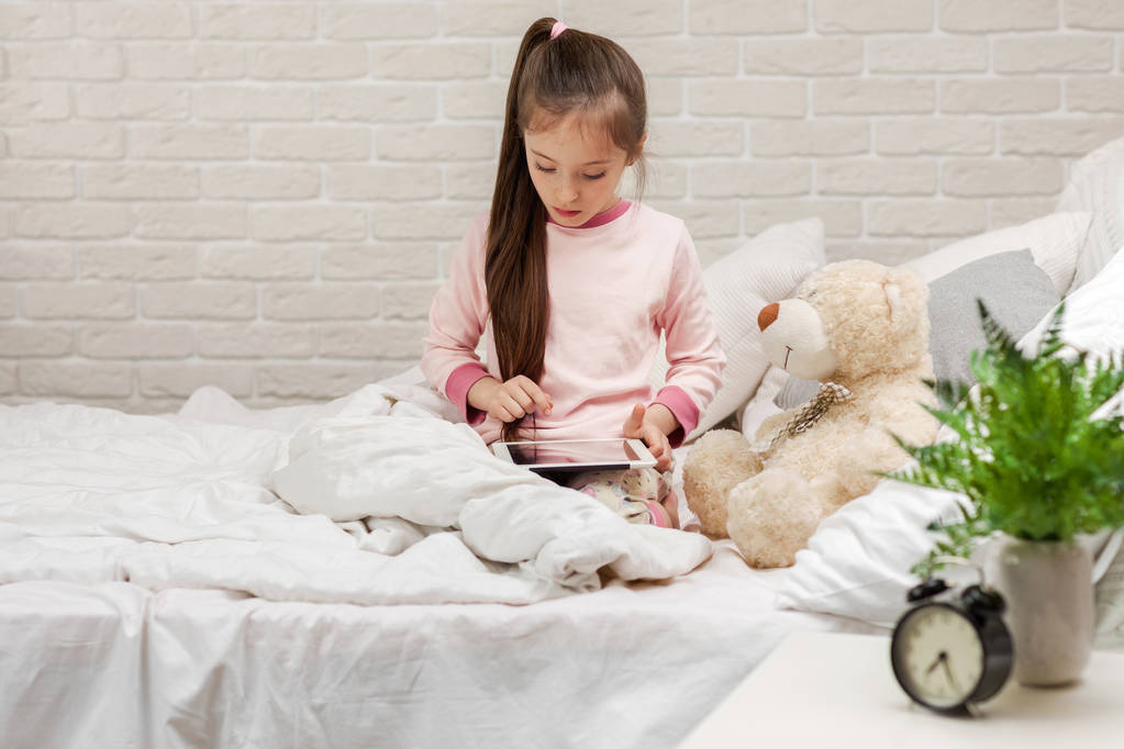 Μικρό κορίτσι βρίσκεται στο κρεβάτι χρησιμοποιεί ψηφιακή ταμπλέτα. - Φωτογραφία, εικόνα
