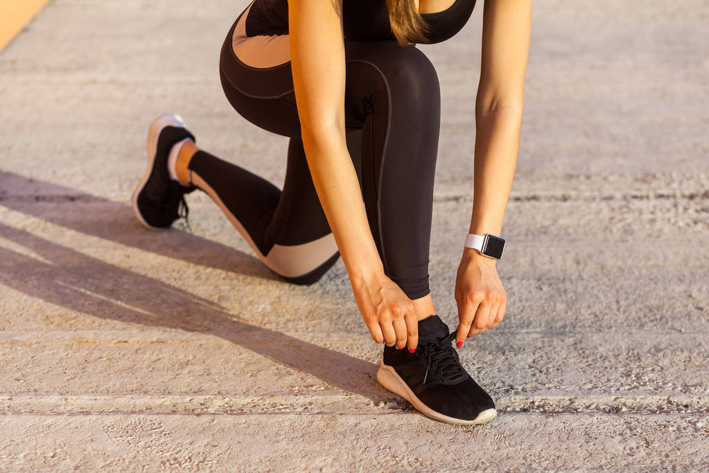 спортивна жінка в чорному спортивному одязі, стоячи на колінах і зав'язуючи шнурки на кросівках на вулиці
  - Фото, зображення