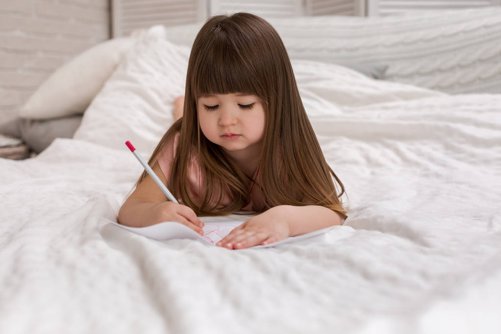 μικρό κορίτσι ζωγραφίζοντας φωτογραφίες ενώ ξαπλωμένοι στο κρεβάτι. - Φωτογραφία, εικόνα