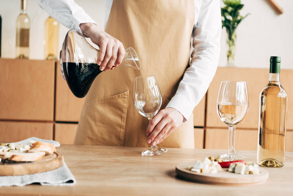 μερική θέα του οινοχόη στην ποδιά κρατώντας κανάτα και χύνοντας κρασί σε γυαλί κρασιού - Φωτογραφία, εικόνα