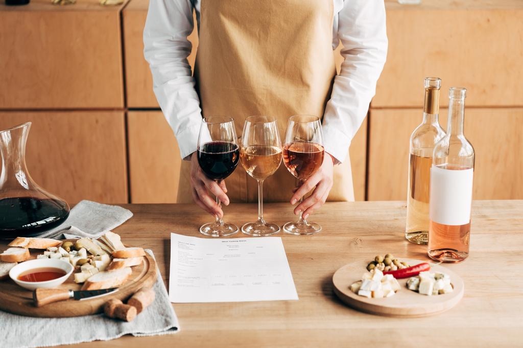 vista recortada de sommelier en delantal sosteniendo copas de vino cerca de mesa de madera
 - Foto, Imagen