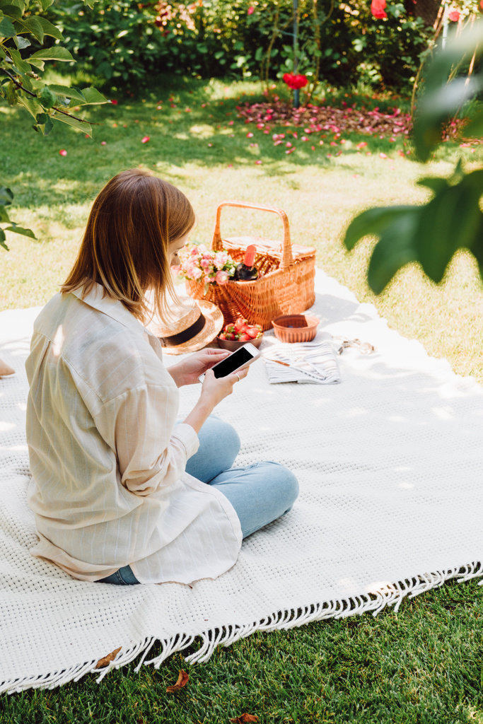 блондинка сидит на одеяле в саду и пикник в солнечный день, используя смартфон
 - Фото, изображение