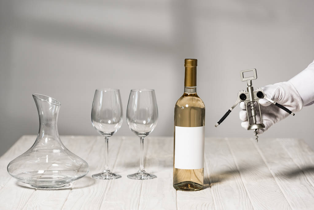 cropped view of waiter in white glove holding corkscrew near table with bottle of wine, wine glasses and jug  - Photo, Image