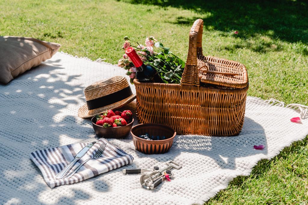 плетеная корзина с розами и вином на белом одеяле возле соломенной шляпы, столовые приборы на салфетке и ягоды в солнечный день в саду
 - Фото, изображение