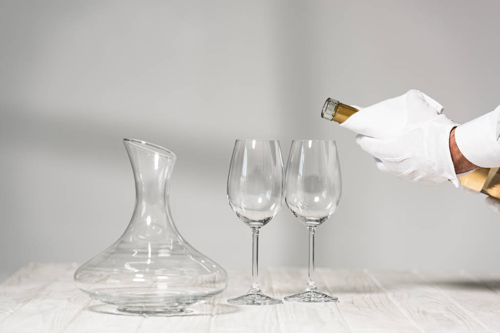 bijgesneden weergave van water in witte handschoen houden fles wijn in de buurt van wijn glazen op houten tafel - Foto, afbeelding