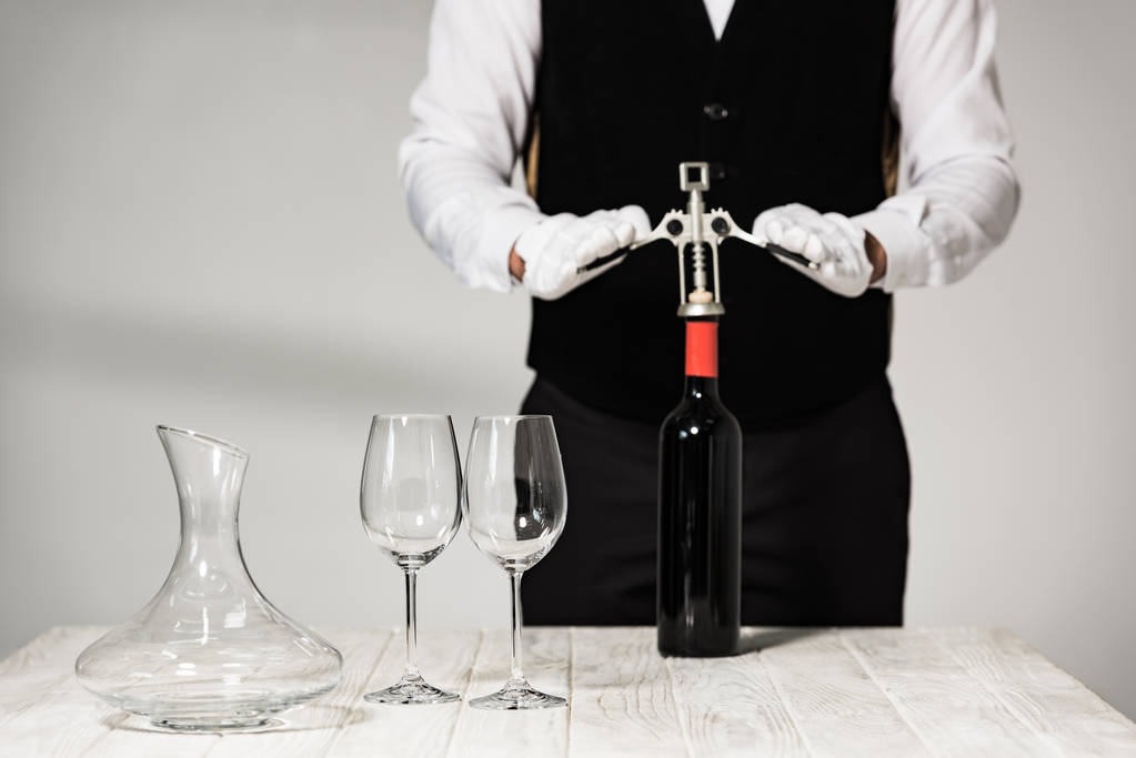 částečný pohled na číšníka v bílých rukavicích otevírající láhev vína s korkšroubem v restauraci - Fotografie, Obrázek