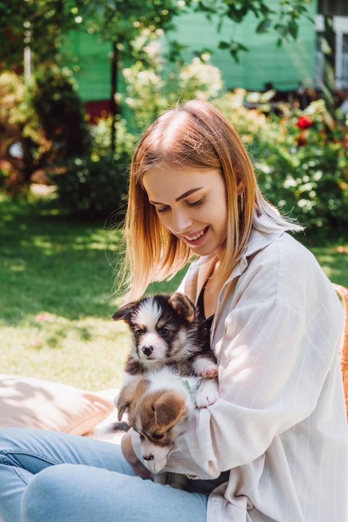happy blonde girl sitting on blanket in garden with adorable puppies at sunny day - Photo, Image