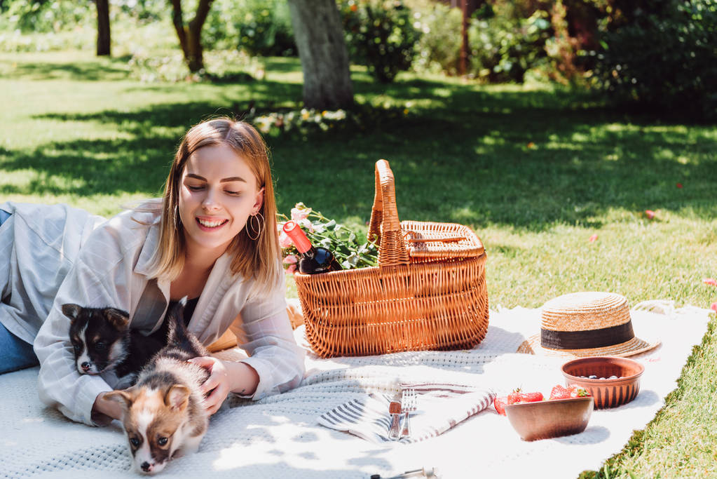 uśmiechnięta blond dziewczyna siedzi na kocu w ogrodzie i mając piknik ze szczeniakami w słoneczny dzień - Zdjęcie, obraz