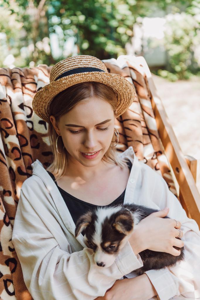 sonrisa rubia chica en paja sombrero celebración cachorro mientras sentado en cubierta silla en jardín
 - Foto, imagen