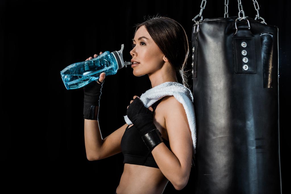 atraktivní dívka pitnou vodu ze sportovní láhve a stojící poblíž děrovacího pytle izolovaného na černém  - Fotografie, Obrázek