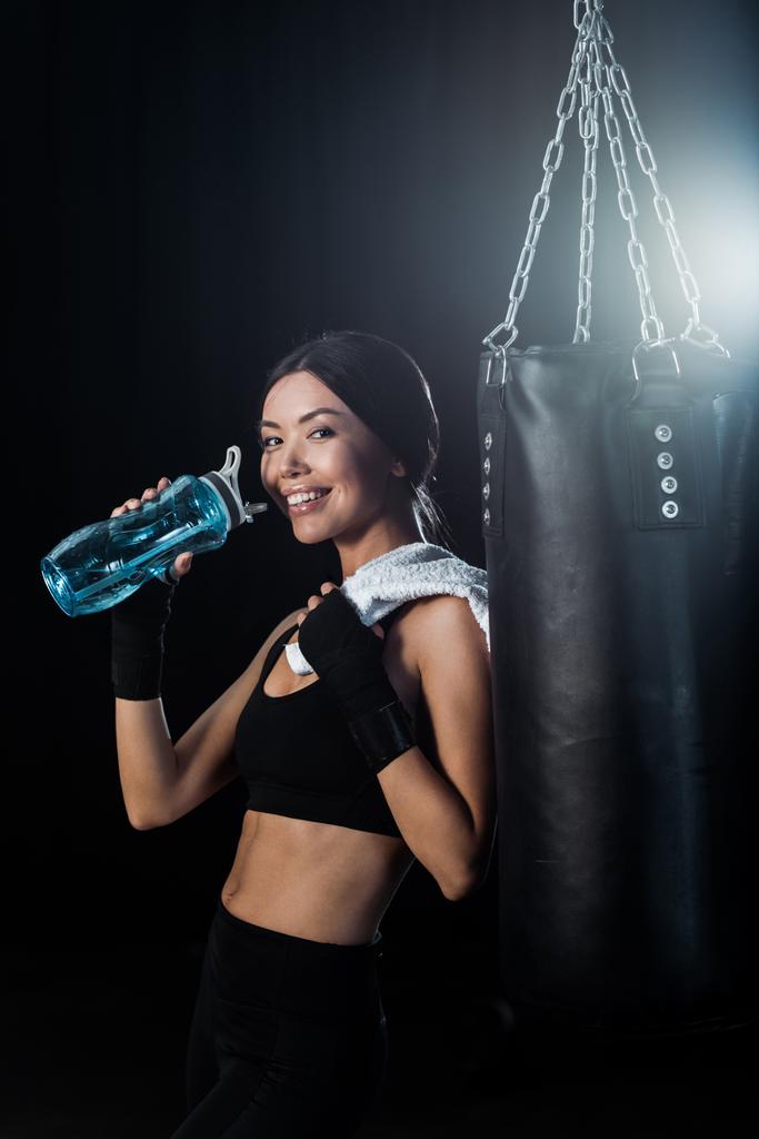 счастливая девушка, держащая спортивную бутылку и стоя рядом с боксерской грушей изолированы на черный
  - Фото, изображение