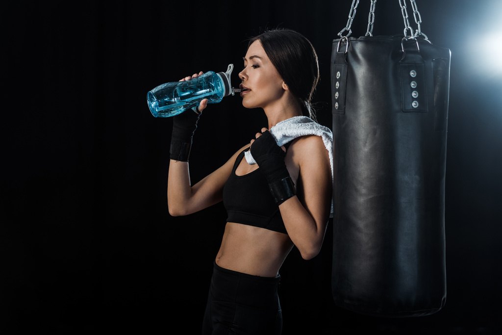 девушка пьет воду из спортивной бутылки и стоит рядом с боксерской грушей изолированы на черный
  - Фото, изображение