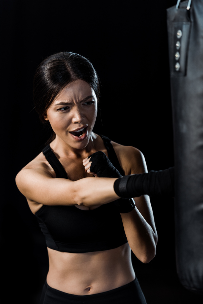 selective focus of emotional sportswoman boxing near punching bag isolated on black  - Photo, Image