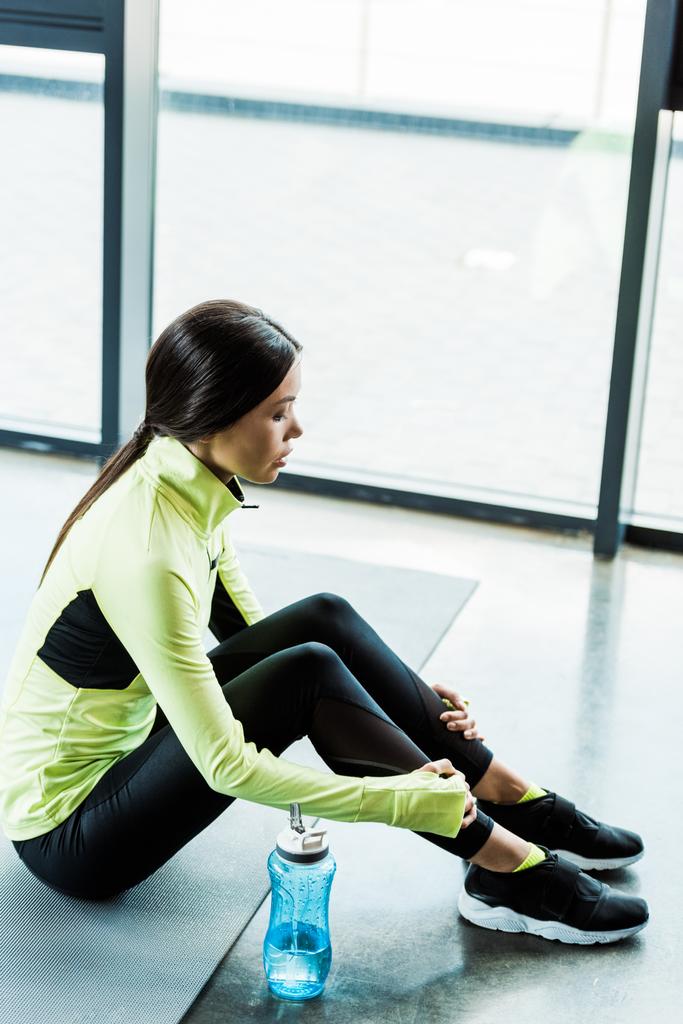 aantrekkelijk meisje in sportkleding zittend op fitness mat in de buurt van sport fles  - Foto, afbeelding