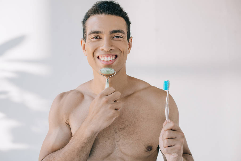 όμορφος αναμεμειγμένη φυλή άνθρωπος μασάζ πρόσωπο με νεφρίτη ρολό και κρατώντας οδοντόβουρτσα, ενώ κοιτάζοντας την κάμερα - Φωτογραφία, εικόνα