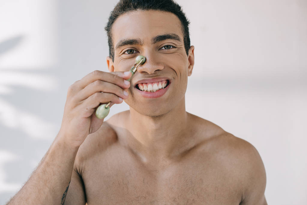 muscular mixed race man massaging nose with stone jade roller and smiling while looking at camera - Photo, Image