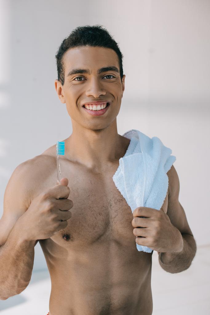 svalnatý muž s modrým ručníkem na rameni a přidržením zubního kartáčku při pohledu na fotoaparát - Fotografie, Obrázek