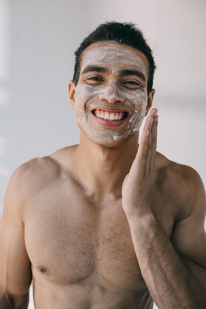 muscoloso uomo di razza mista che applica crema cosmetica sul viso con mano e sorridente mentre guarda la fotocamera
 - Foto, immagini