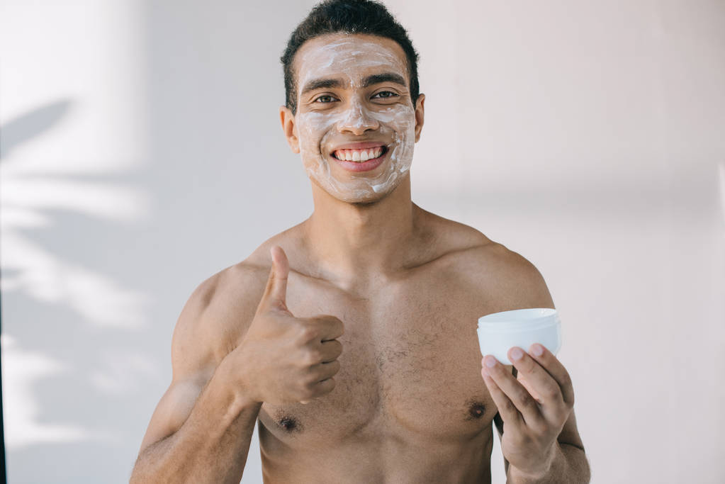 handsome mixed race man with cosmetic cream on face holding container and showing thumb up while smiling and looing at camera - Photo, Image
