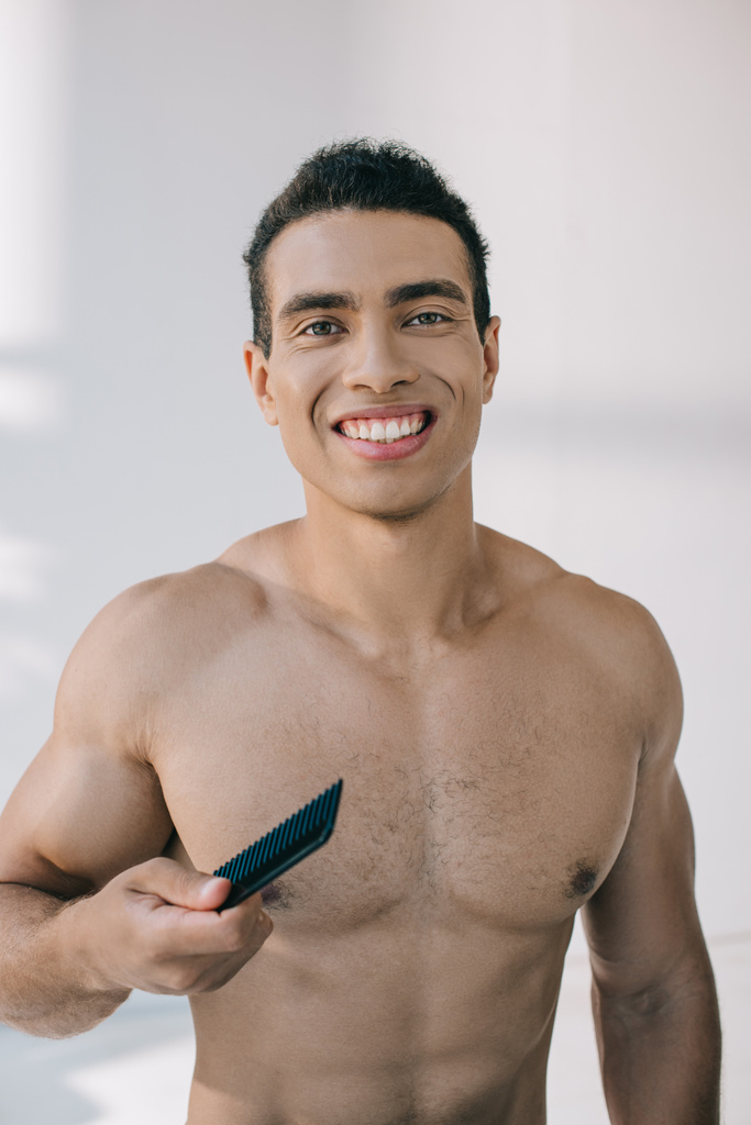 handsome mixed race man holding hairbrush and smiling while looking at camera - Photo, Image
