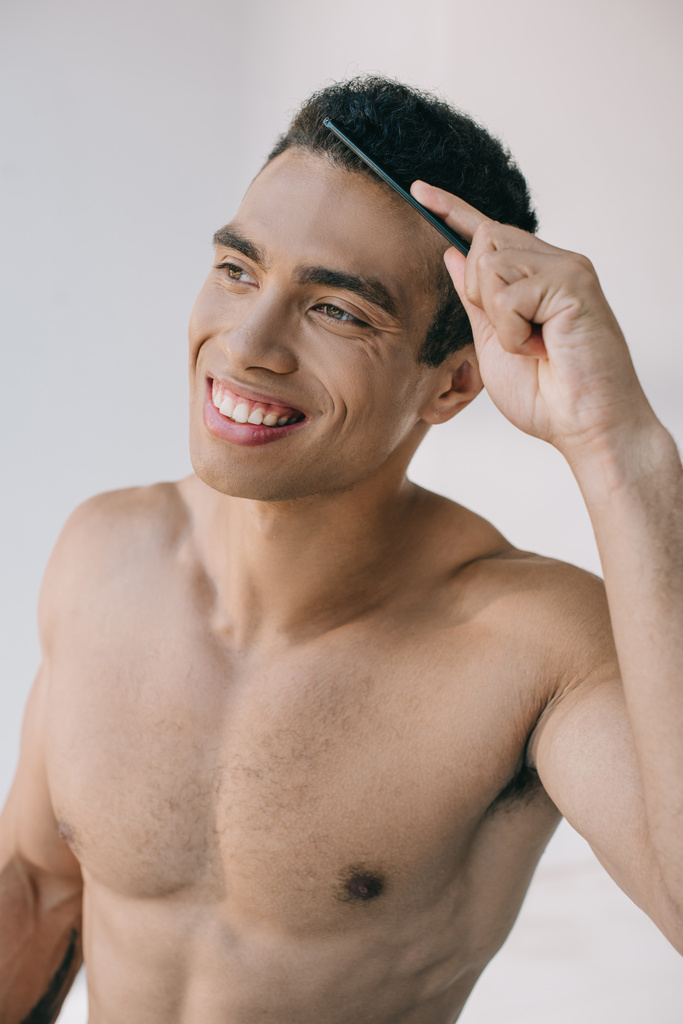 handsome mixed race man combing hair with hairbrush and smiling while looking away - Photo, Image