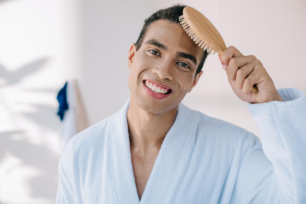 handsome young man in bathrobe combing hair with hairbrush while amiling and looking at camera - Photo, Image