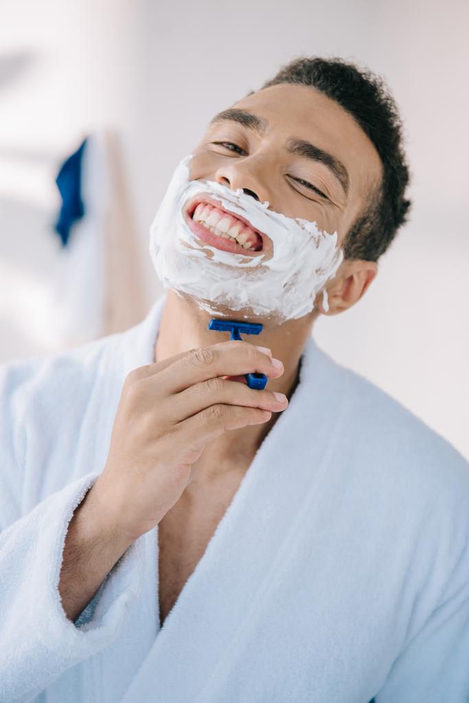 portré lövés jóképű ember borotválkozás arcát borotvával, miközben mosolyogva és néztem kamera - Fotó, kép