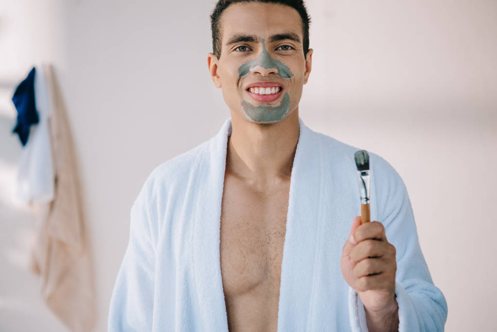 bonito homem segurando escova enquanto de pé em roupão de banho e com máscara de barro no rosto
 - Foto, Imagem