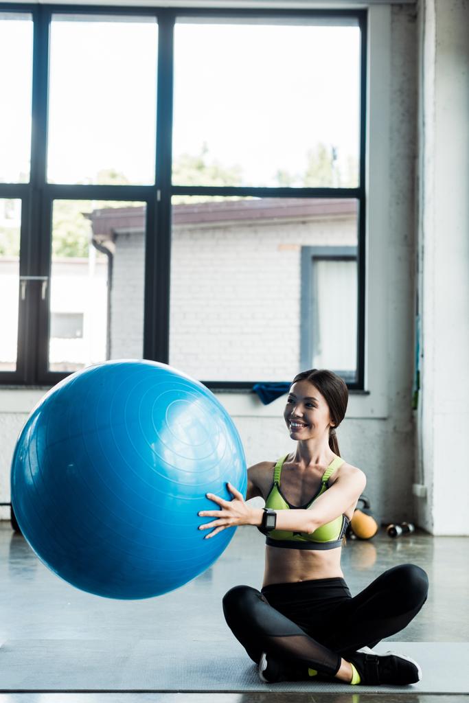 menina alegre segurando bola de fitness azul enquanto sentado no tapete de fitness
  - Foto, Imagem