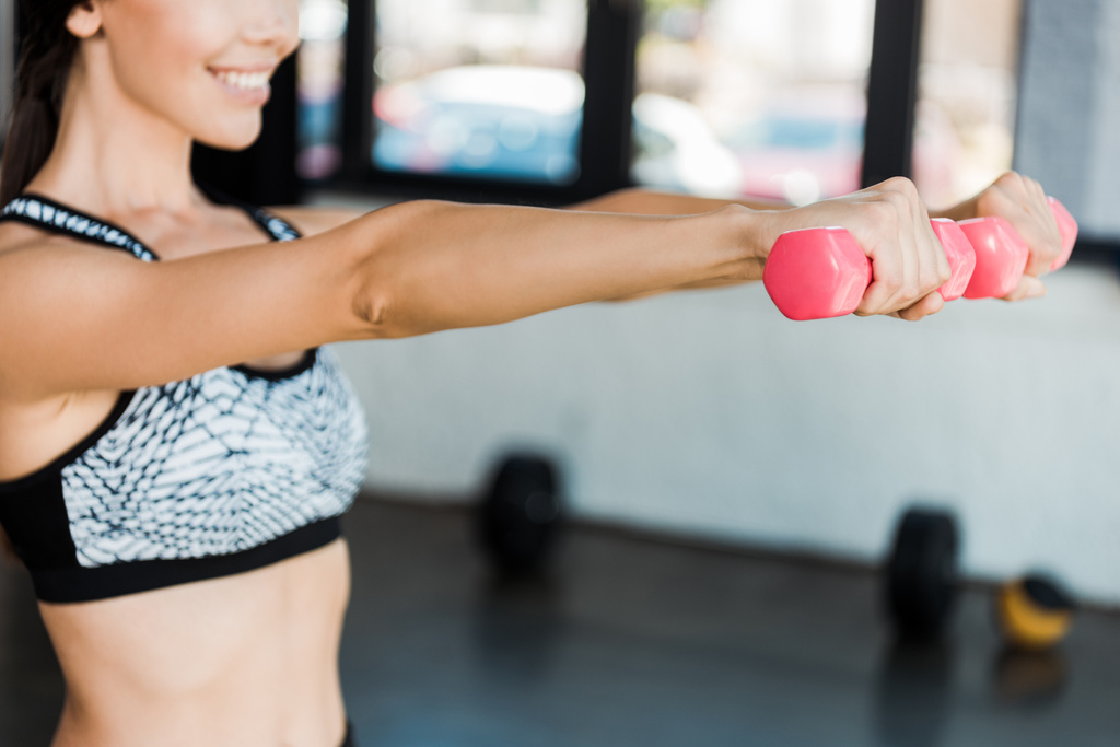 cropped view of happy girl holding pink dumbbells while exercising in gym  - Photo, Image