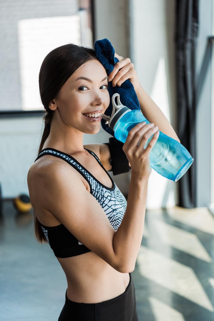 vrolijke jonge vrouw die sport fles vasthoudt en zweet in het sportcentrum veegt  - Foto, afbeelding