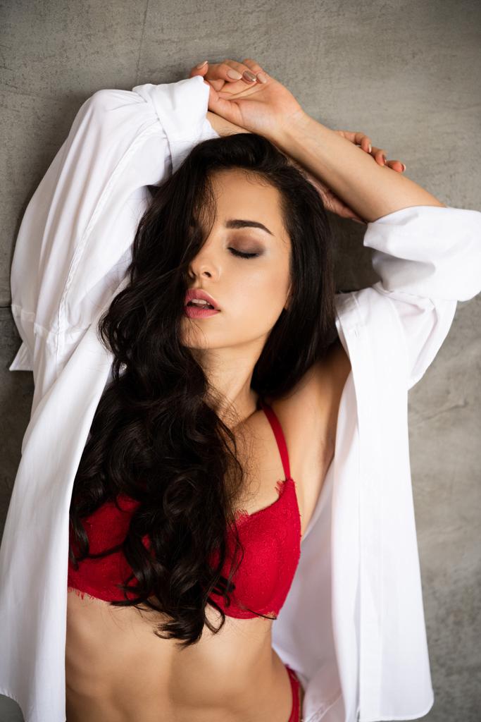 retrato de chica sexy en ropa interior roja y camisa blanca apoyada en la pared gris con la mano sobre la cabeza y con los ojos cerrados
 - Foto, imagen