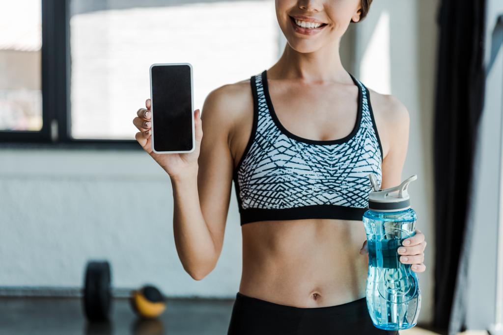 cropped view of cheerful girl holding sport bottle and smartphone with blank screen  - Photo, Image