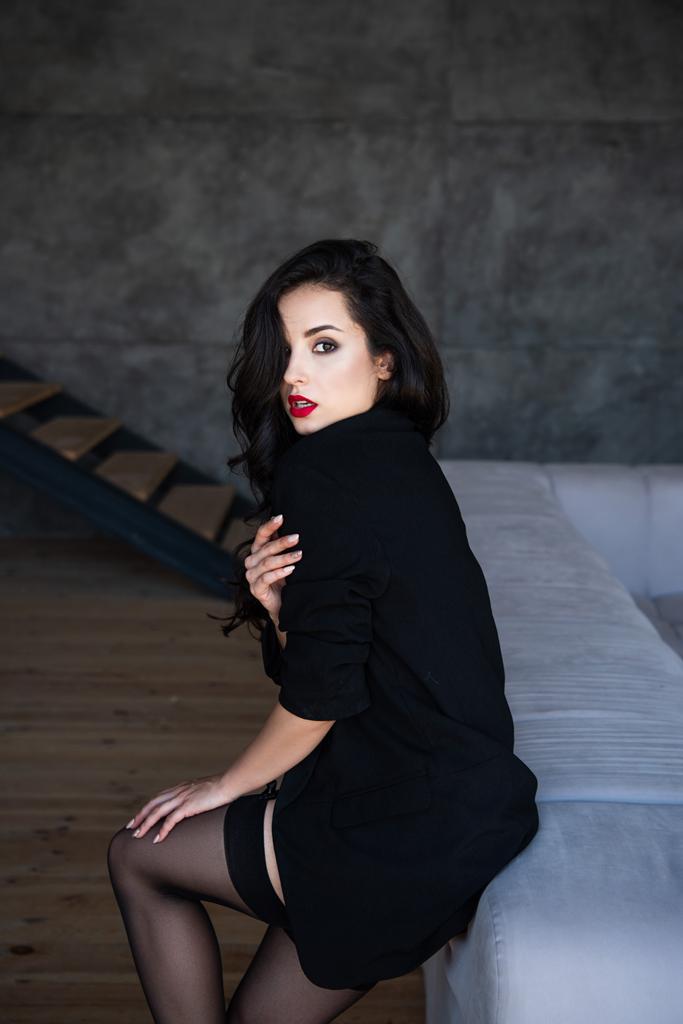 chica sexy con chaqueta negra sentado en el sofá y mirando a la cámara
 - Foto, Imagen