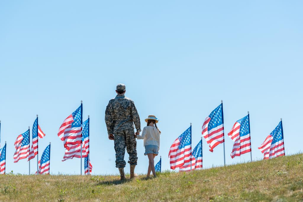 back view of man in military uniform holding hands with daughter near american flags  - Photo, Image