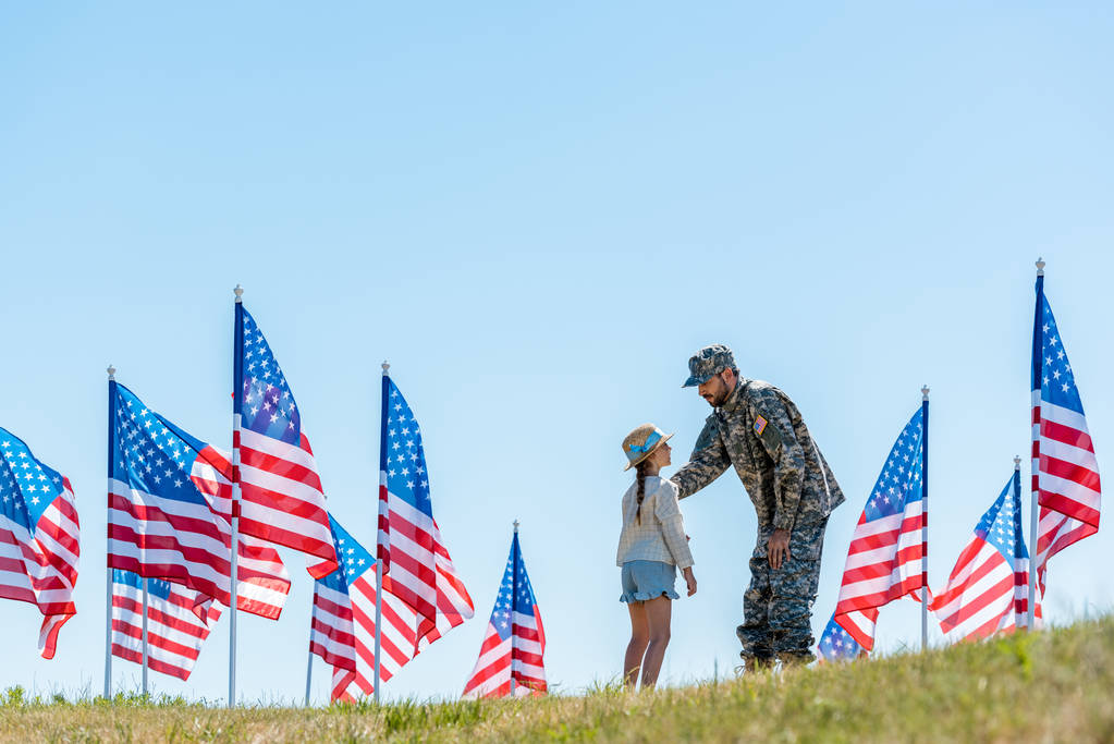 Selektiver Fokus eines Mannes in Militäruniform, der seine Tochter in der Nähe amerikanischer Flaggen betrachtet  - Foto, Bild