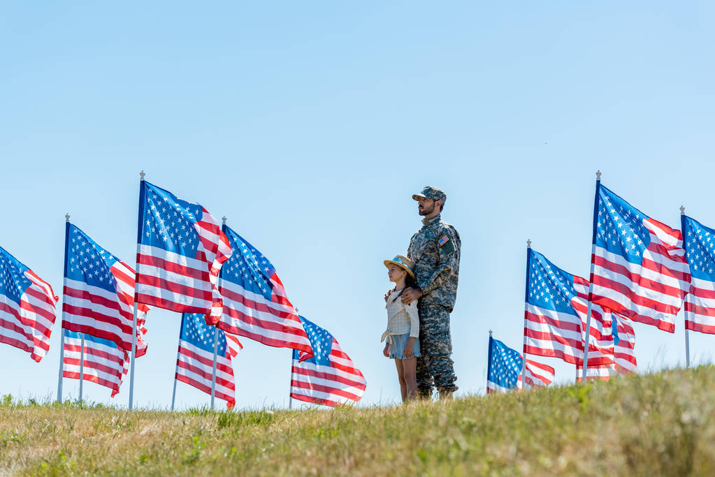 επιλεκτική εστίαση του πατέρα σε στρατιωτική ομοιόμορφη στέκεται με χαριτωμένο παιδί κοντά αμερικανικές σημαίες  - Φωτογραφία, εικόνα