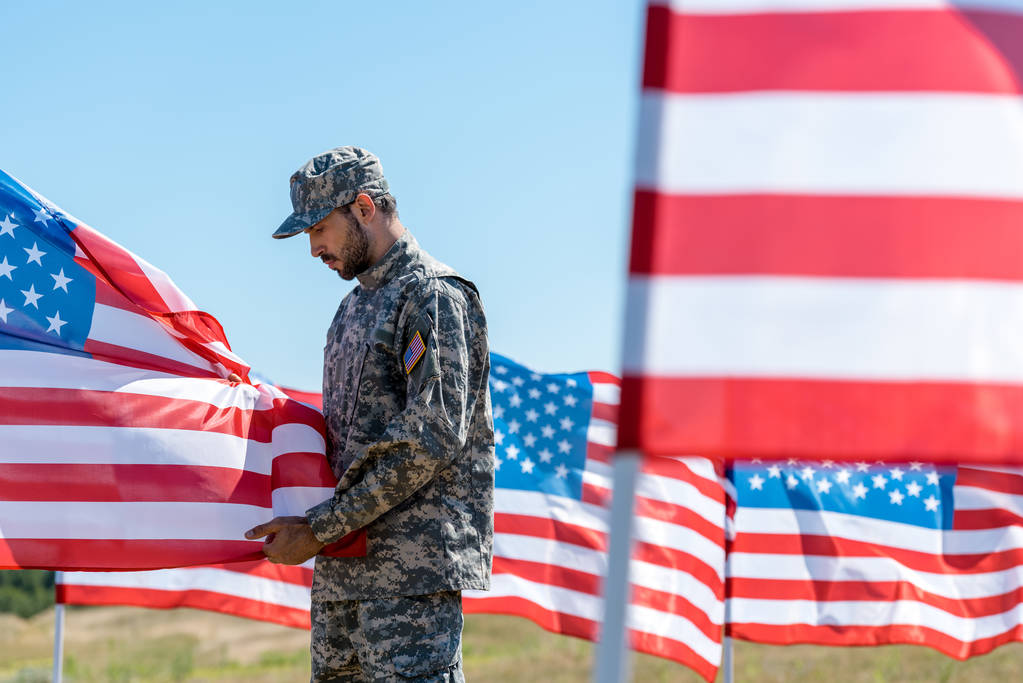 селективный фокус человека в военной форме и кепке стоя и касаясь американского флага
  - Фото, изображение