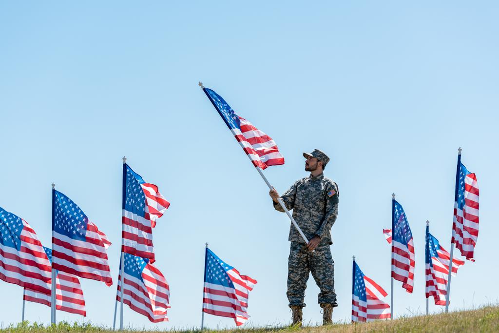 εκλεκτική εστίαση του όμορφου στρατιώτη με στρατιωτική στολή και καπάκι που κρατά την αμερικάνικη σημαία  - Φωτογραφία, εικόνα
