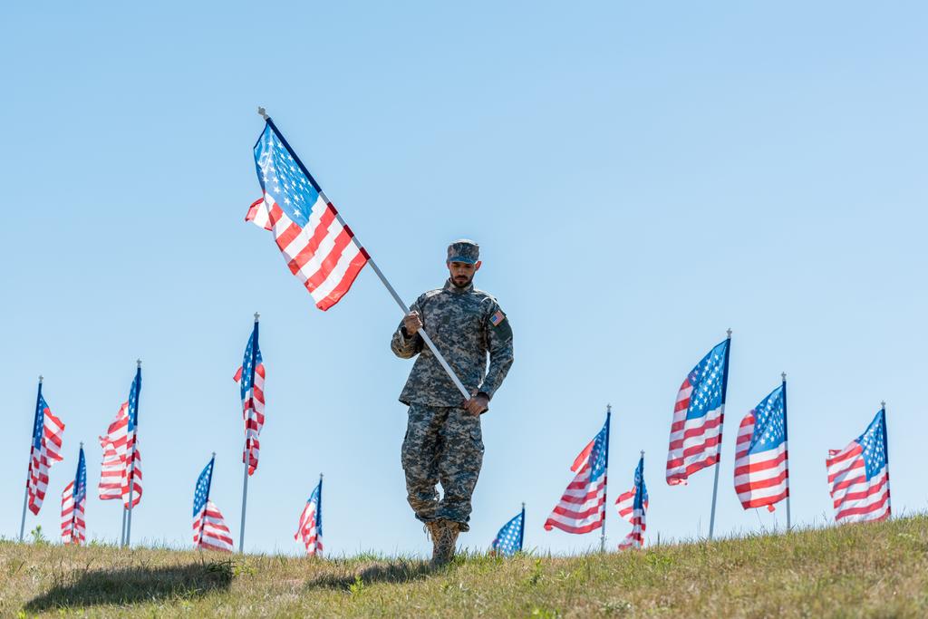 schöner Soldat in Militäruniform und Mütze mit amerikanischer Flagge, während er im Gras steht  - Foto, Bild