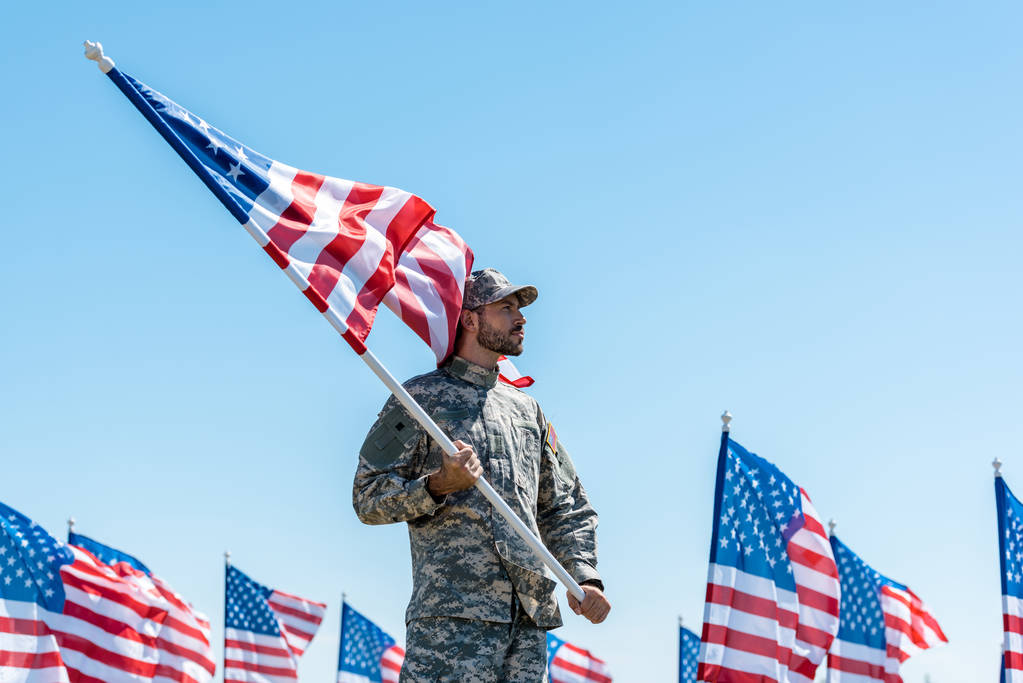 knappe man in militair uniform en Cap Holding Amerikaanse vlag terwijl staande tegen blauwe hemel  - Foto, afbeelding