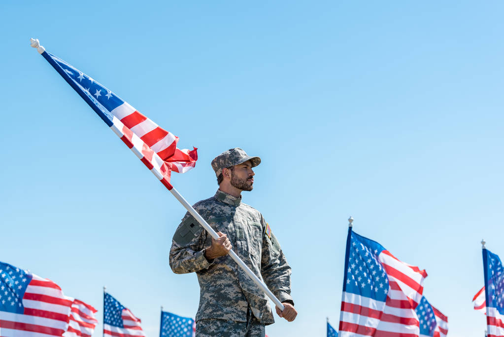 guapo soldado en uniforme militar y gorra sosteniendo bandera americana mientras está de pie contra el cielo azul
  - Foto, imagen