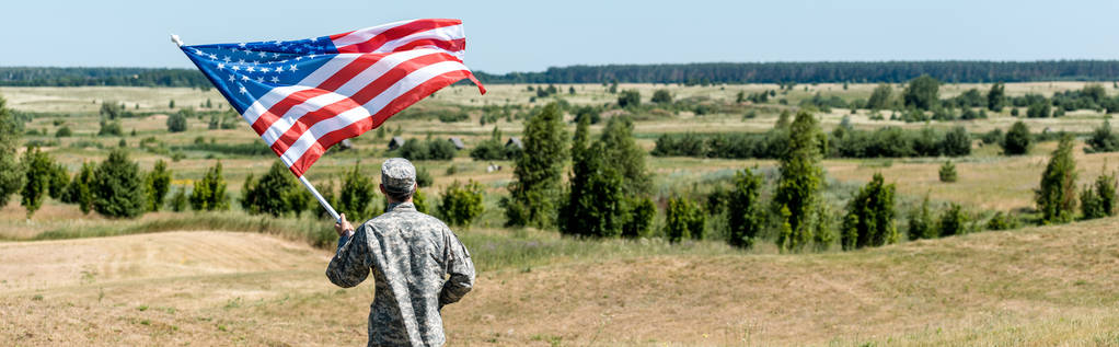 Panoramaaufnahme eines Militärmannes, der neben Bäumen steht und die amerikanische Flagge hält  - Foto, Bild