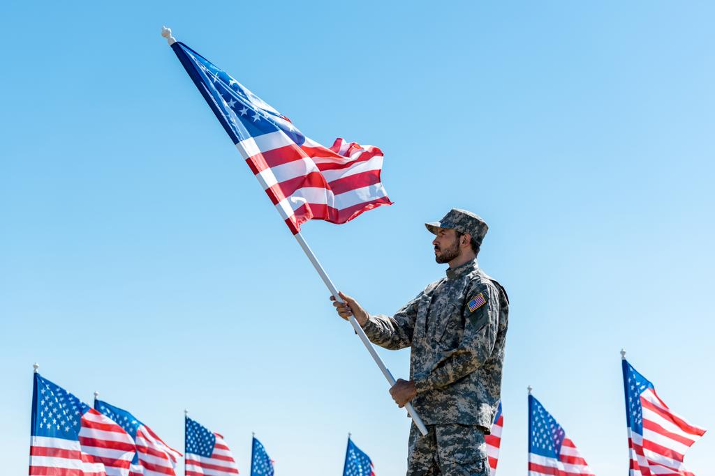 guapo militar en uniforme y gorra sosteniendo bandera americana mientras está de pie contra el cielo azul
  - Foto, imagen