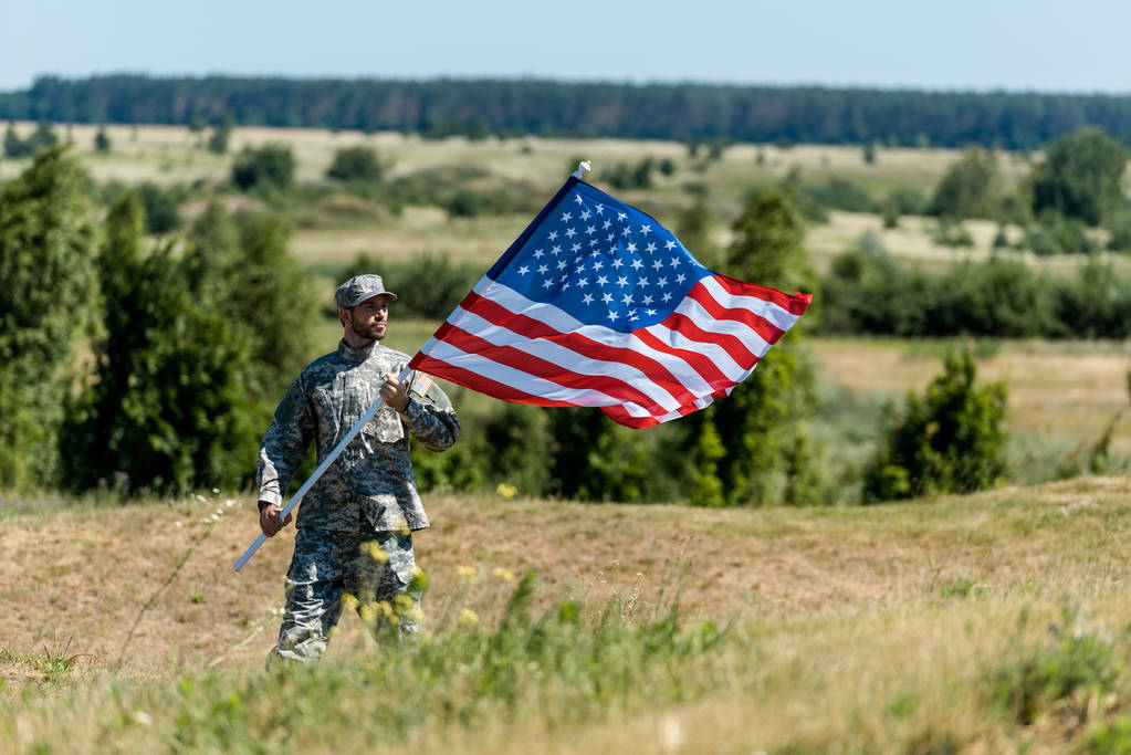 εκλεκτική εστίαση του όμορφου στρατιωτικού άντρα με στολή και καπέλο που κρατά την αμερικάνικη σημαία το καλοκαίρι  - Φωτογραφία, εικόνα