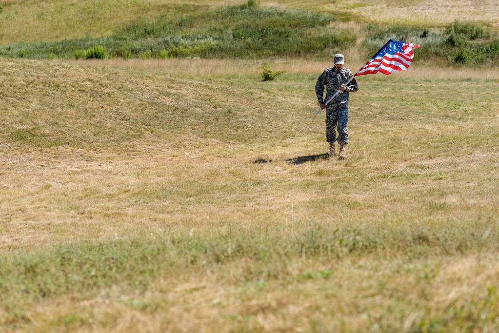 selectieve focus van knappe soldaat in uniform wandelen en het houden van Amerikaanse vlag in de zomer  - Foto, afbeelding